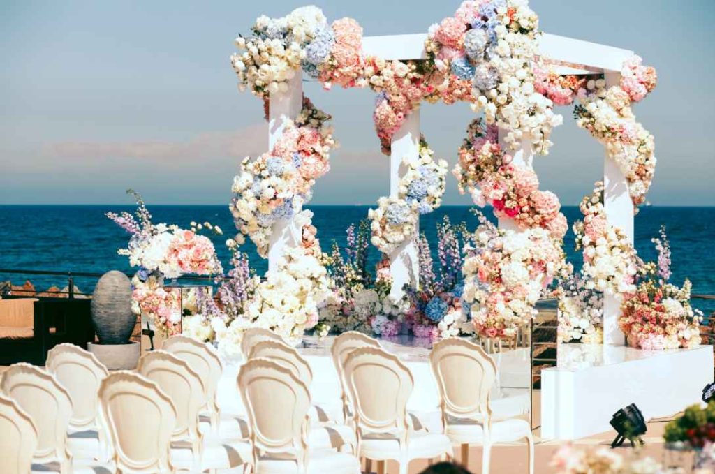 Arche florale blanche et chaises élégantes dressées face à la mer pour organiser une cérémonie de mariage sur la plage.