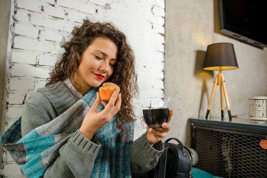 Une femme aux cheveux bouclés savoure une boisson chaude et un petit gâteau dans un intérieur chaleureux.